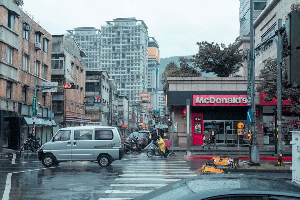 Rainy urban crosswalk scene with traffic, pedestrians, and high-rise buildings including a McDonald's in a downtown area.